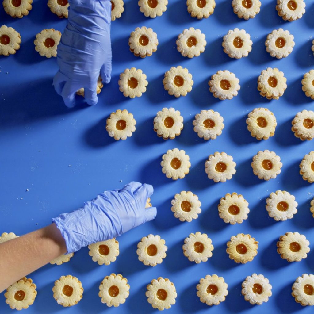 A vibrant overhead view of neatly arranged cookies with a jam center on a blue conveyor belt, featuring a worker's gloved hand picking up one of the cookies.